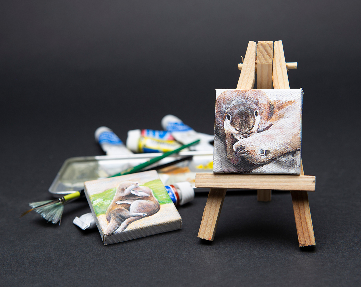 Mini-canvas painting of otters on a mini-easel with a second painting and art supplies.